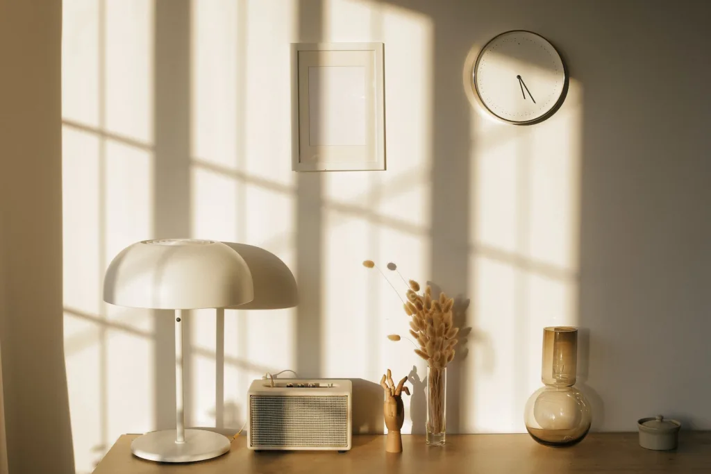 Light room with retro radio and decorative vases with dry plants on desk near wall with clock and window shadow in sunlight 11society.com