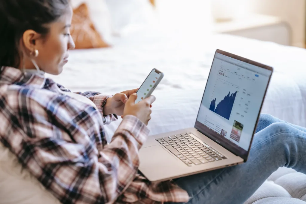 Investing in ETFs - Side view of crop ethnic female dealer using trading app on cellphone against netbook with graph on screen in house