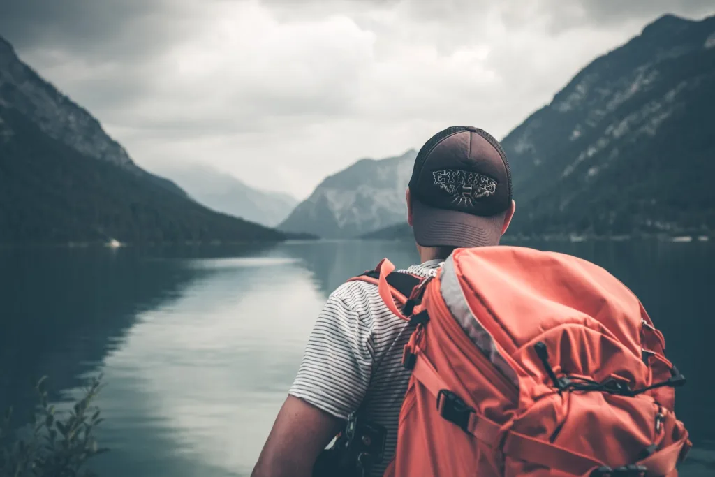 Travel Hacks - man with red hiking backpack facing body of water and mountains at daytime