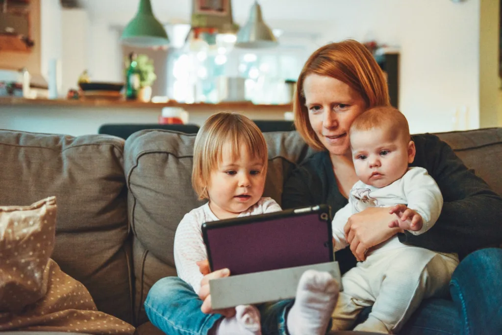 Teaching kids about money - two babies and woman sitting on sofa while holding baby and watching on tablet
